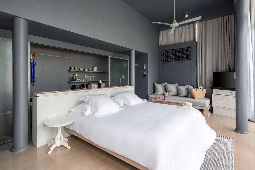 Modern bedroom with a large white bed, gray walls, a small side table, open shelves, a sitting area with a sofa and coffee table, a TV, floor-to-ceiling curtains, and a ceiling fan.