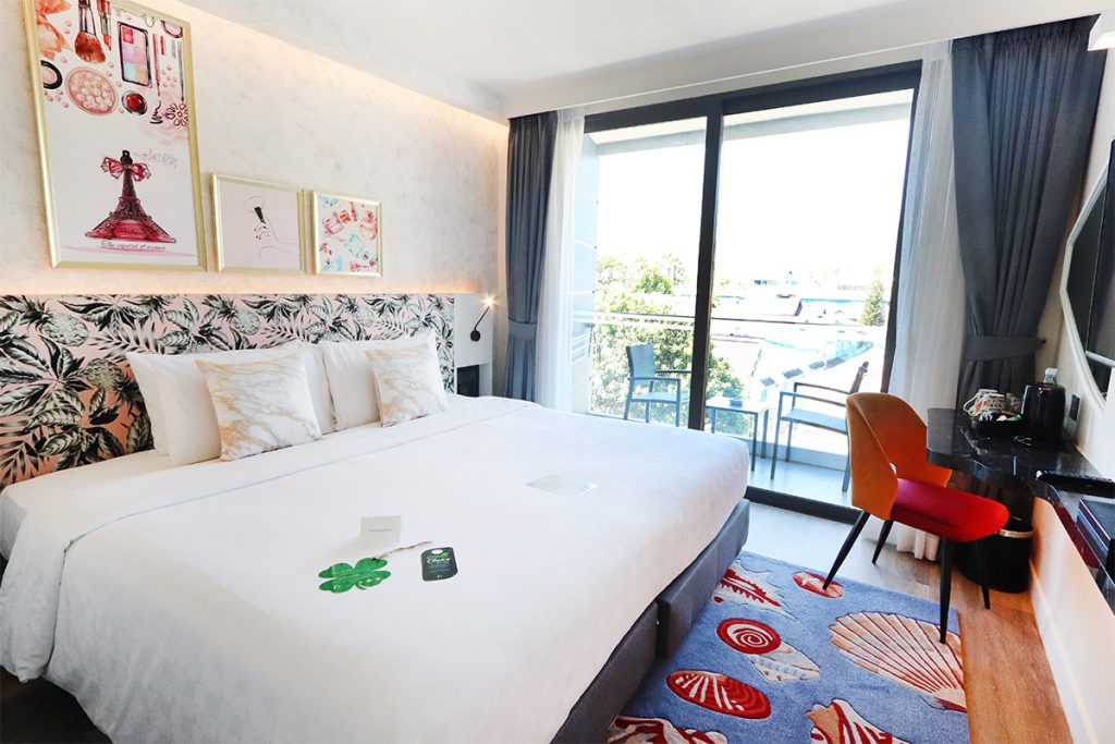 A modern hotel room with a large bed, colorful artwork above the headboard, a balcony with outdoor seating, and a desk with a red chair. The room is bright with natural light from sliding glass doors.
