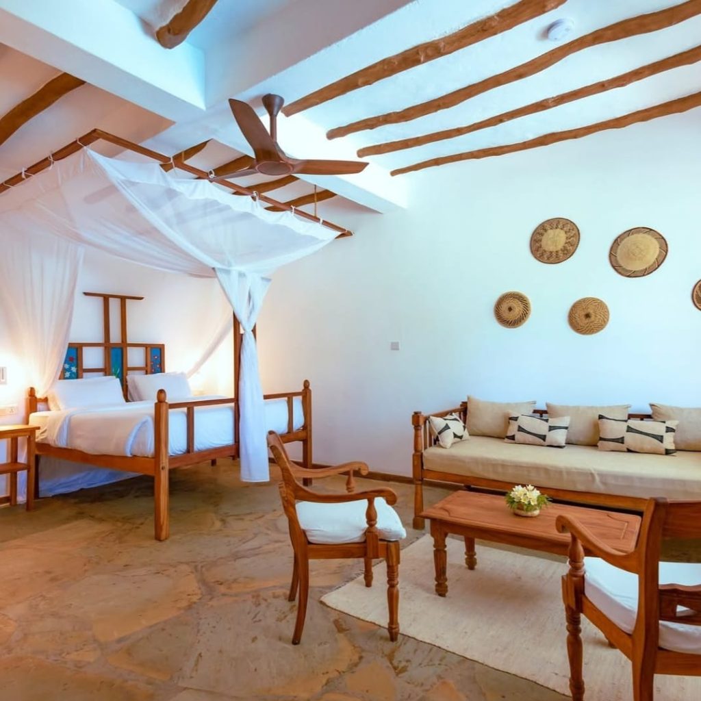 A bright, airy bedroom features a wooden canopy bed with white netting, a beige sofa with throw pillows, wooden chairs around a coffee table, and woven baskets hanging on the white wall as decor.