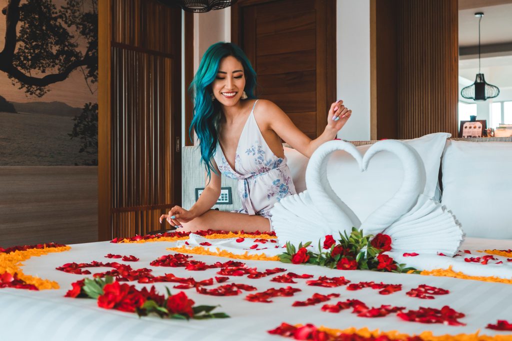 A woman with blue hair in a light dress smiles while sitting on a bed decorated with rose petals and towel swans forming a heart in a bright, modern room.