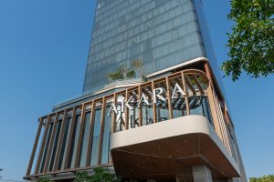 Modern high-rise building with a glass facade and the name AKARA displayed prominently above the entrance, viewed from a low angle against a clear blue sky. Trees partially frame the image.