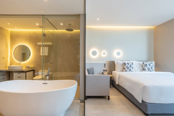 Modern hotel room with a large bed, armchair, and a bathroom featuring a freestanding bathtub, round mirror, and glass shower enclosure. Soft lighting creates a clean, inviting atmosphere.