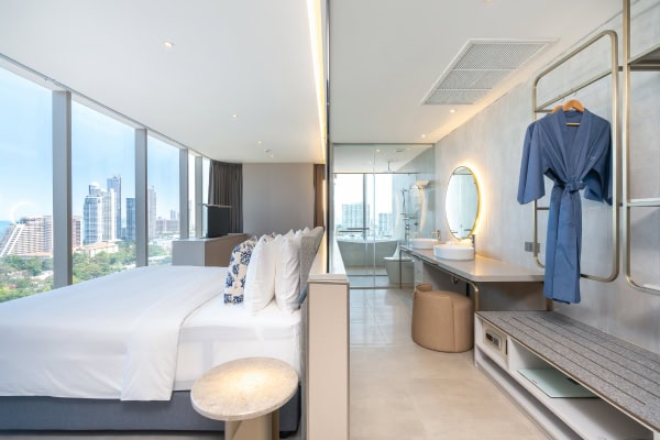 A modern hotel room with large windows showing a city skyline, a neatly made bed, a bathroom area with a round mirror, and a blue robe hanging on the wall. The decor is bright and minimalist.