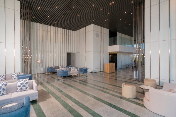 Modern hotel lobby with high ceilings, striped flooring, white walls, blue and beige seating areas, decorative pillows, metal accents, and a clock on the wall above a small wooden reception desk.