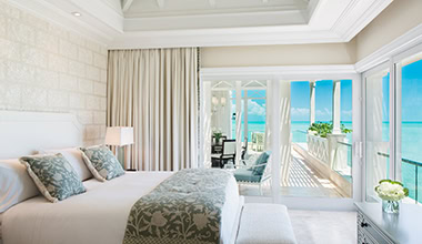 A bright, elegant bedroom with a large bed, pastel bedding, sheer curtains, and sliding glass doors opening to a balcony overlooking a turquoise ocean and clear blue sky.