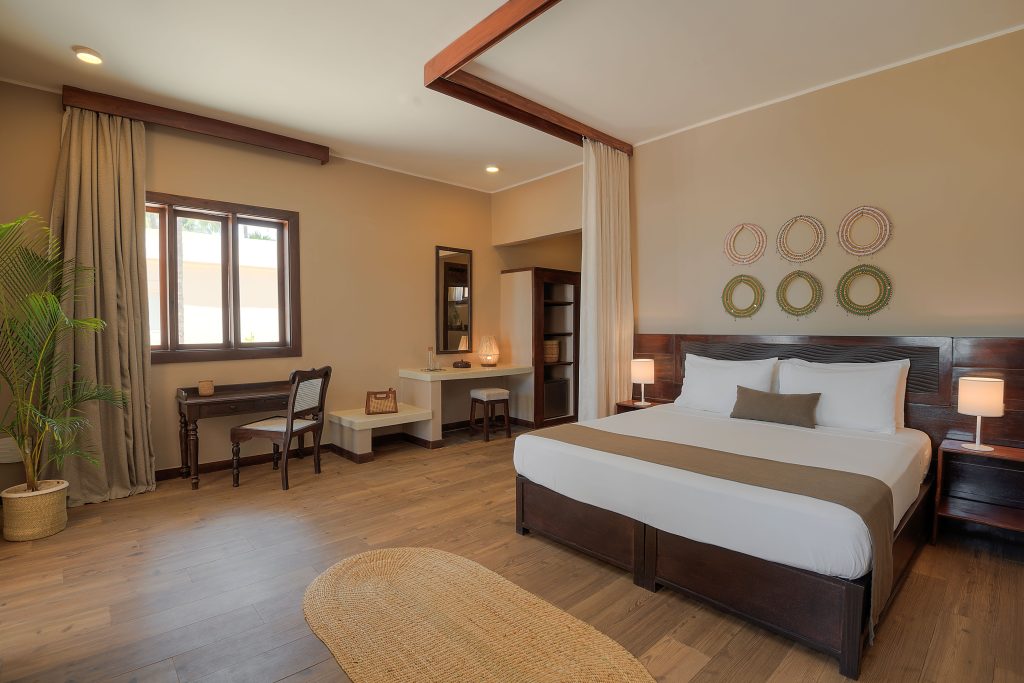 A spacious hotel room with a large bed, wooden floor, desk, chair, potted plant, two windows with curtains, soft lighting, wall art above the bed, and a cozy, minimalist decor style.