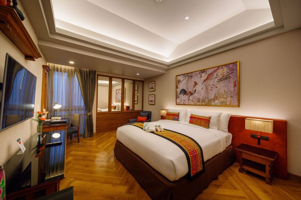 A cozy hotel room with a large bed, colorful pillows and runners, wooden flooring, a desk with a chair, wall art depicting animals, and warm lighting creating a welcoming atmosphere.