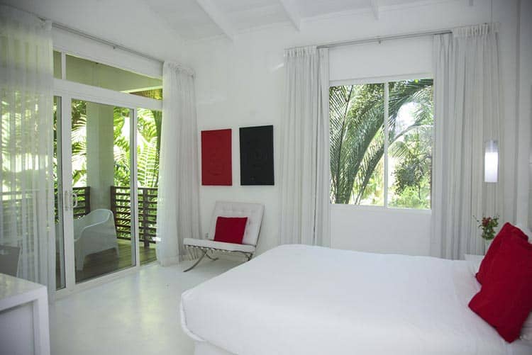 Bright, modern bedroom with white walls, bed, and chair. Red pillows and wall art provide contrast. Large windows and glass doors reveal lush greenery outside, letting in natural light.