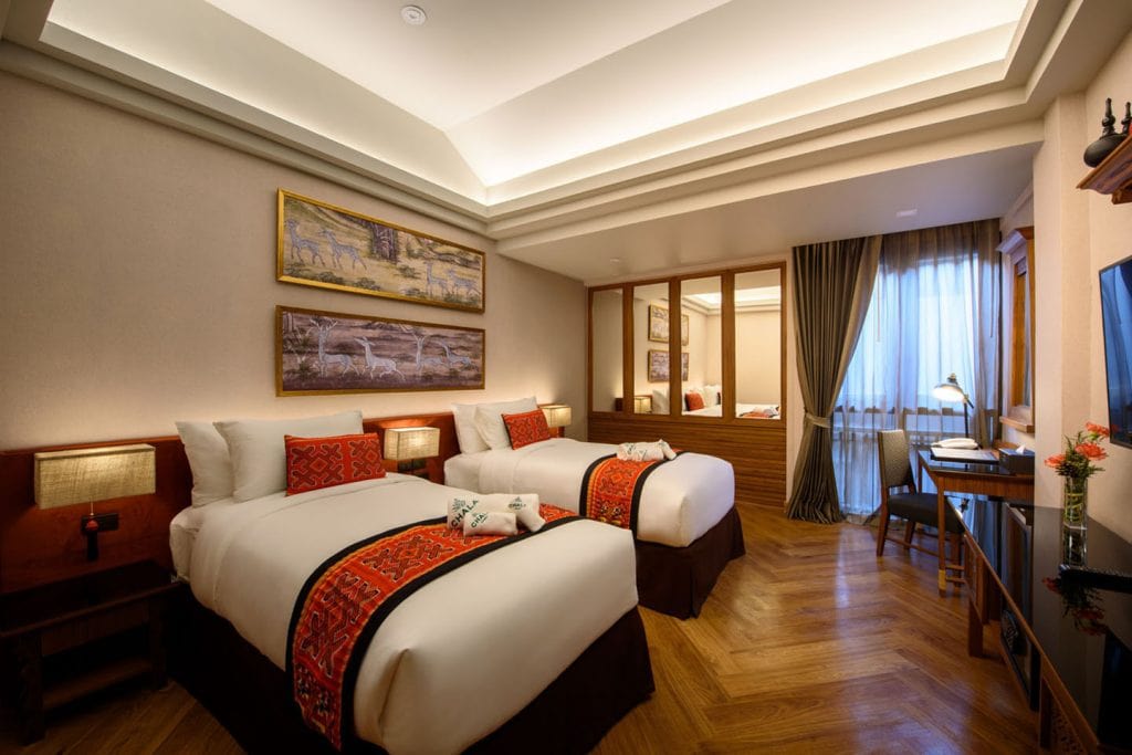 A hotel room with two neatly made twin beds, red decorative pillows and runners, wooden floors, a desk with a chair by a large window, warm lighting, and paintings of animals on the wall.