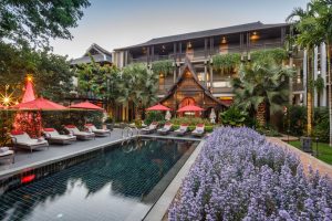 A luxurious hotel courtyard with a swimming pool, surrounded by purple flowers, palm trees, and sun loungers with red umbrellas. A decorated Christmas tree and lush greenery accent the elegant multi-story building in the background.
