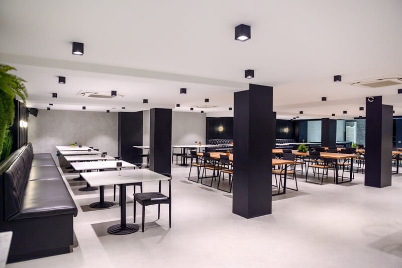 A modern, spacious restaurant with white walls and ceiling, black pillars, and minimalist black and wooden tables and chairs. Green plants decorate some tables, and long black benches line one wall. The restaurant is empty.