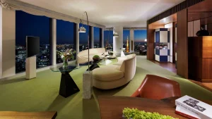 A modern hotel suite with curved sofas, a glass coffee table, floor-to-ceiling windows showing a city skyline at night, and stylish decor including flowers and books on tables.