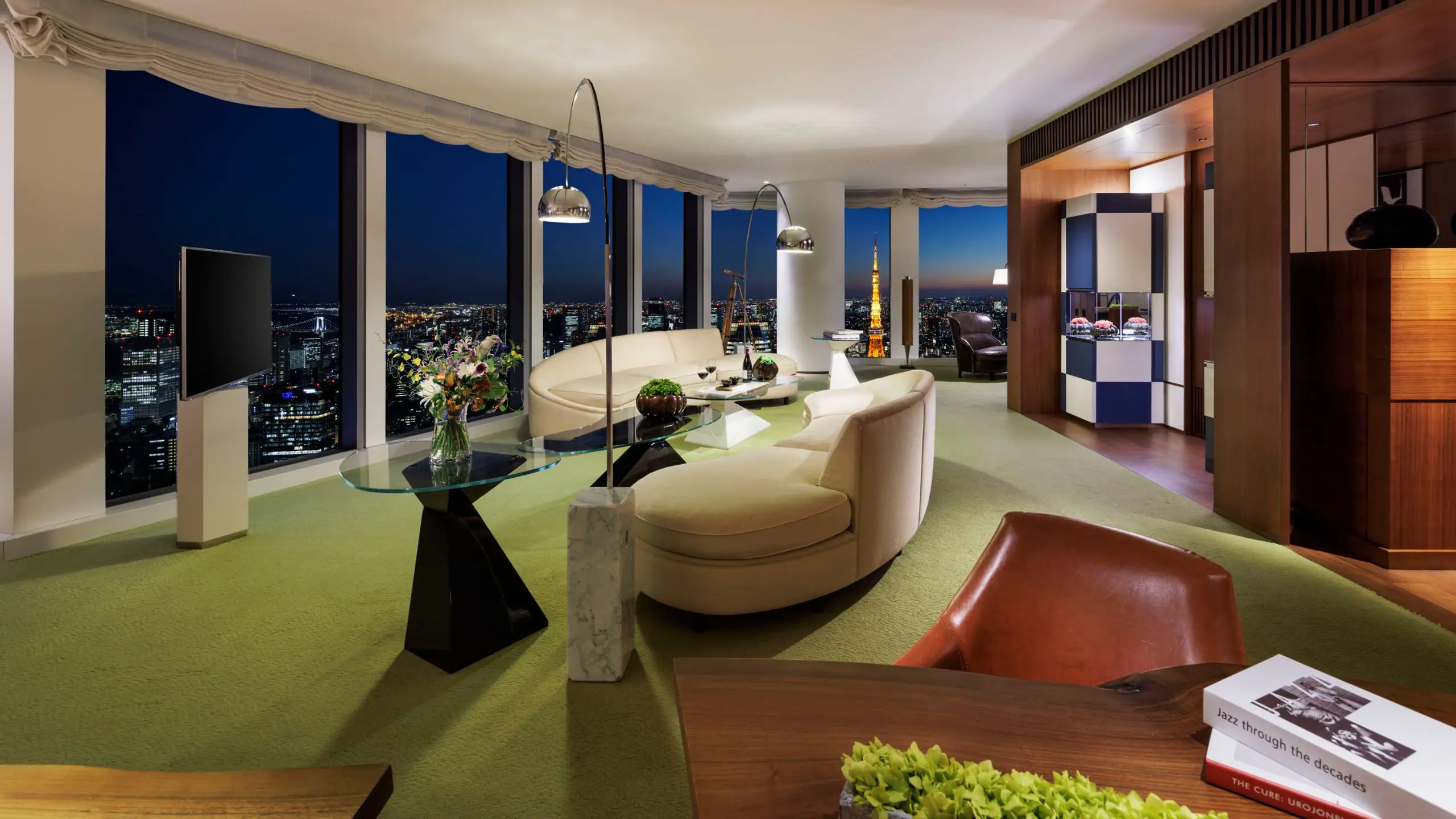 A modern hotel suite with curved sofas, a glass coffee table, floor-to-ceiling windows showing a city skyline at night, and stylish decor including flowers and books on tables.