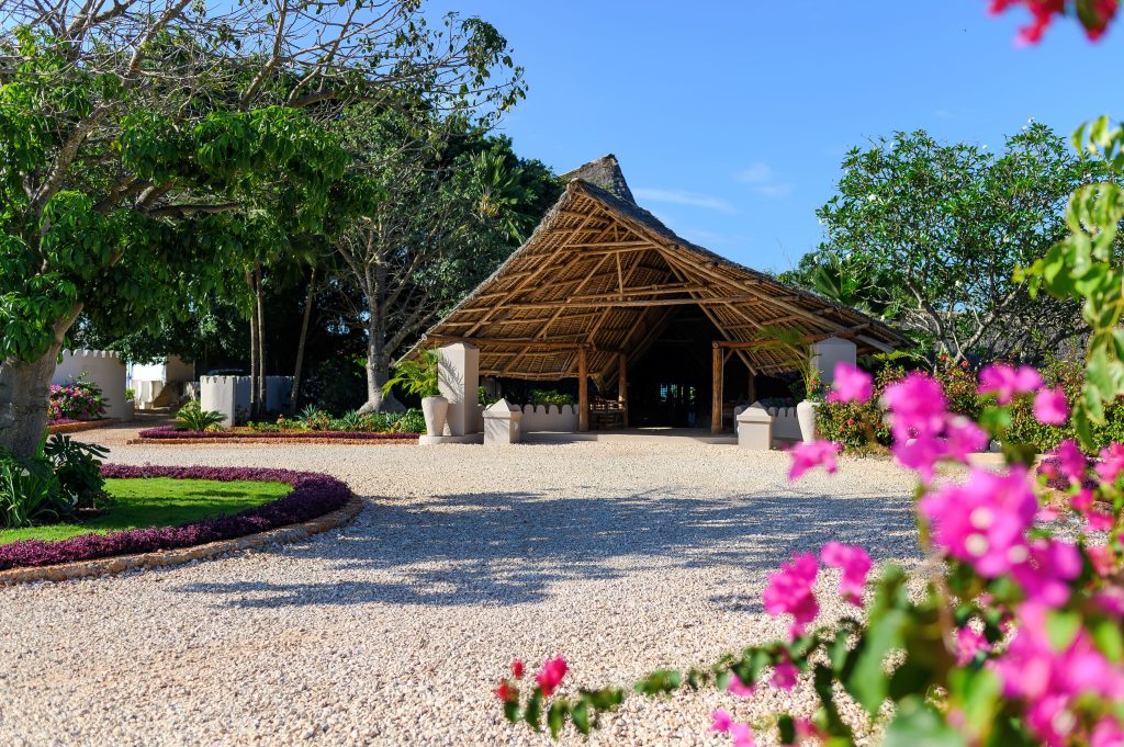 A thatched-roof pavilion stands in a garden with trees, flowering plants, and a gravel pathway under a clear blue sky. Pink flowers are in the foreground.