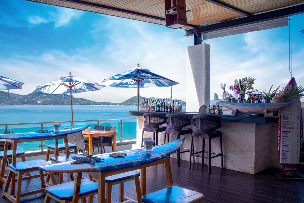 A beachfront bar with surfboard-shaped tables, wicker chairs, and blue umbrellas overlooks a turquoise ocean and distant hills under a bright, partly cloudy sky. Bottles and flowers decorate the bar counter.