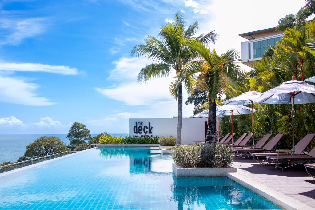 Infinity pool with clear blue water, surrounded by palm trees and lounge chairs with umbrellas, overlooking the ocean under a bright blue sky. A sign reads The Deck Pool Lounge.