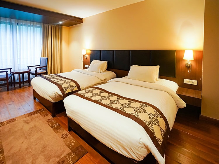 A hotel room with wooden floors, two single beds with white bedding and patterned runners, wall-mounted lamps above the headboards, a window with curtains, and a small seating area with two chairs and a table.