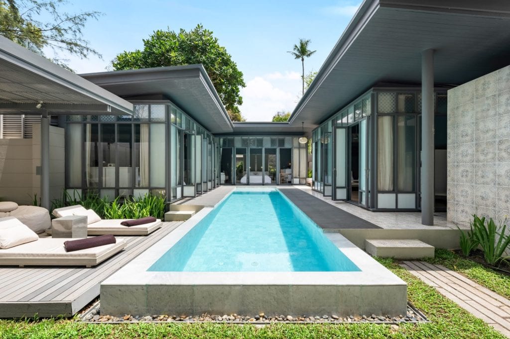Modern villa with large glass windows and a rectangular swimming pool in the center, surrounded by lounge chairs and greenery, under a bright blue sky.