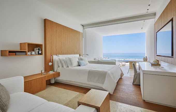 A modern hotel room with a large bed, wooden accents, a sofa, a desk, and a wide window offering a clear view of the ocean and blue sky. The space is bright, minimalist, and elegantly decorated.