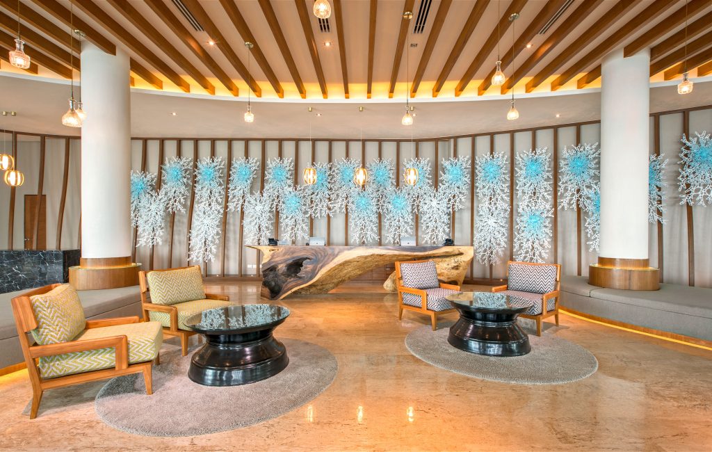 Modern hotel lobby with stylish seating, glass tables, a large sculpted wooden centerpiece, and a decorative wall featuring blue and white abstract art. Warm lighting and wooden accents create an inviting atmosphere.