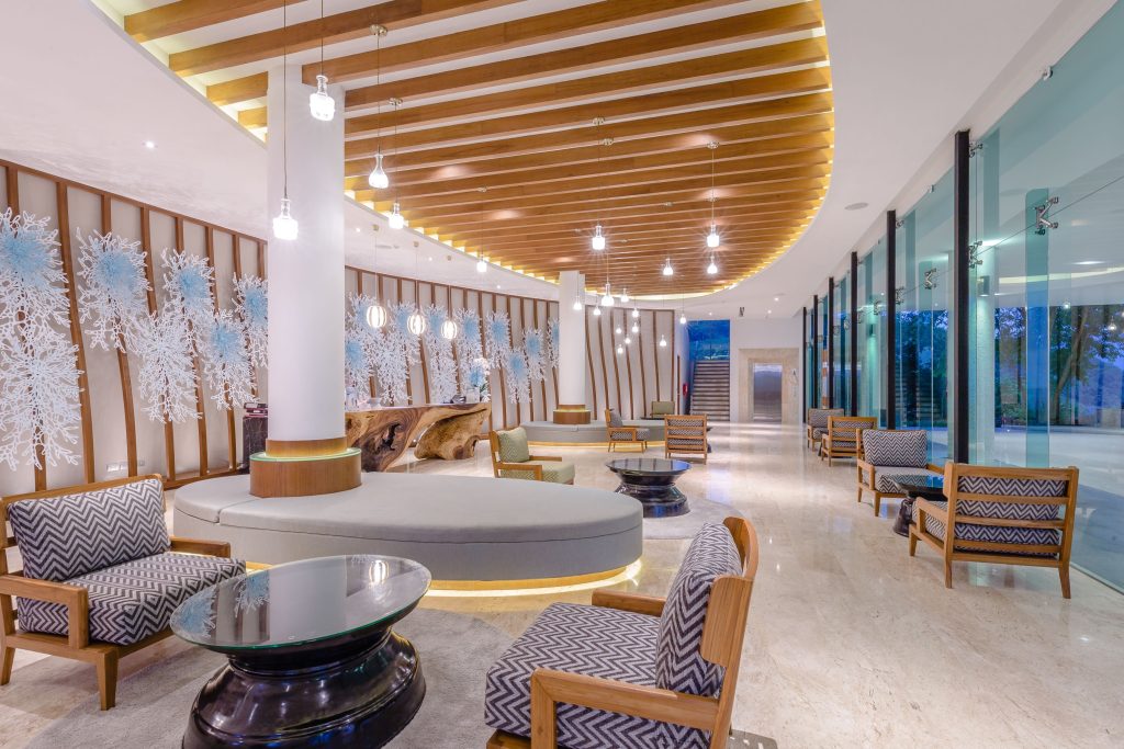 A modern hotel lobby with stylish seating, wooden accents, artistic wall decor, large glass windows, and a glossy marble floor, illuminated by hanging lights.