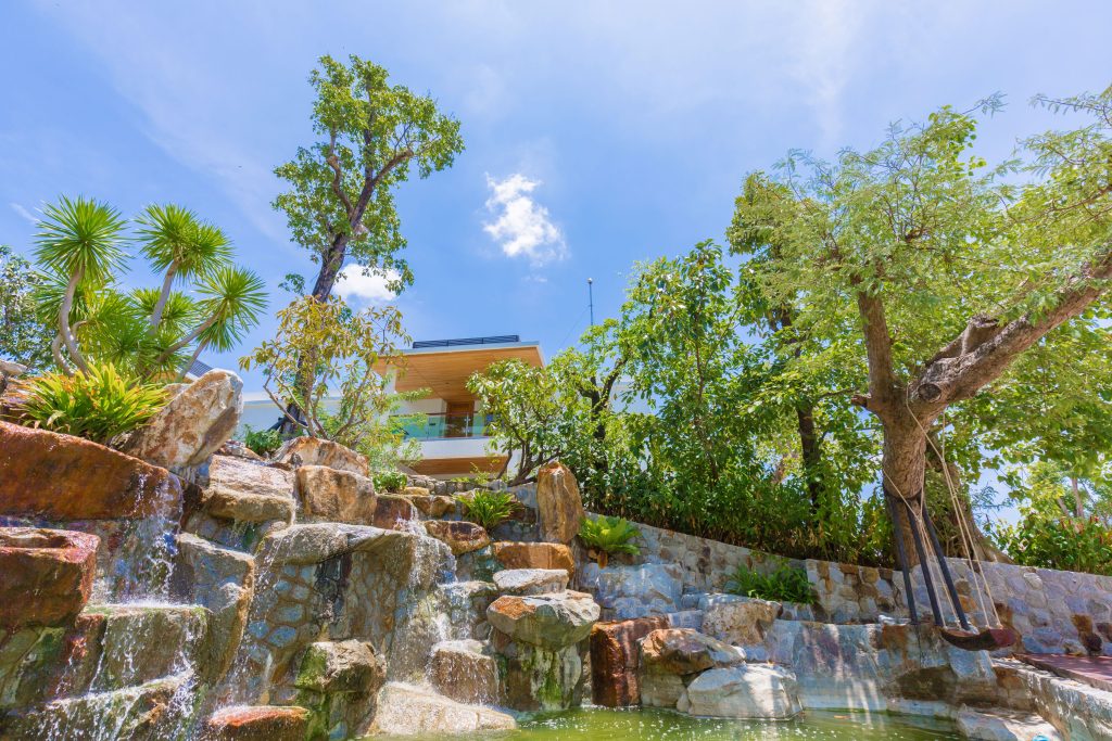 A modern house partially hidden by lush green trees and plants, with a small waterfall flowing over rocks into a pond under a bright blue sky with a few clouds.