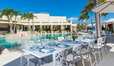 Elegant outdoor dining area set with white tables and chairs beside a turquoise pool, surrounded by palm trees and modern buildings under a clear blue sky.