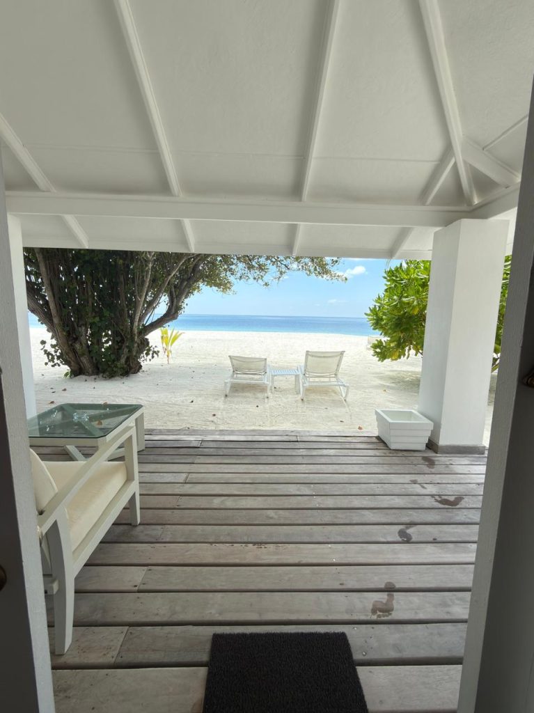A view from a shaded porch with wooden flooring looking out onto a sandy beach. Two white lounge chairs face the calm sea, surrounded by green trees and clear blue sky in the background.