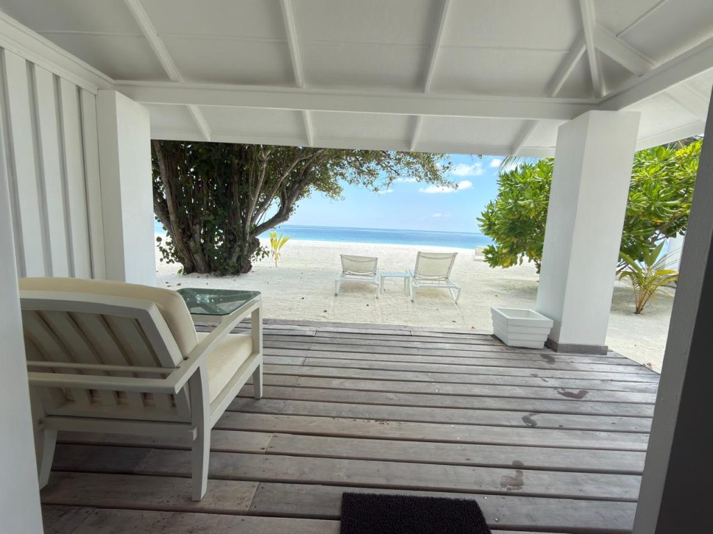 A shaded wooden patio with white furniture overlooks a sandy beach with two lounge chairs facing the calm blue ocean, framed by green trees under a bright sky.