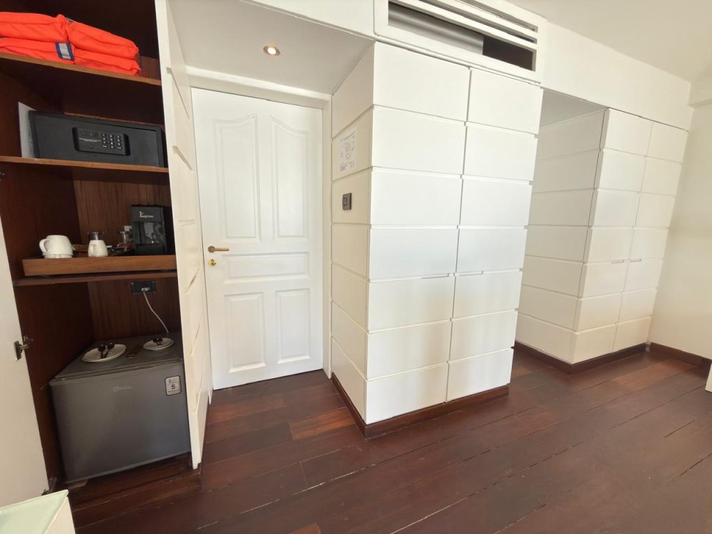 A hotel room corner with wooden floor, white paneled walls, a closed white door, a mini fridge, a shelf with a safe, coffee maker, cups, and a bright orange life jacket on the top shelf.