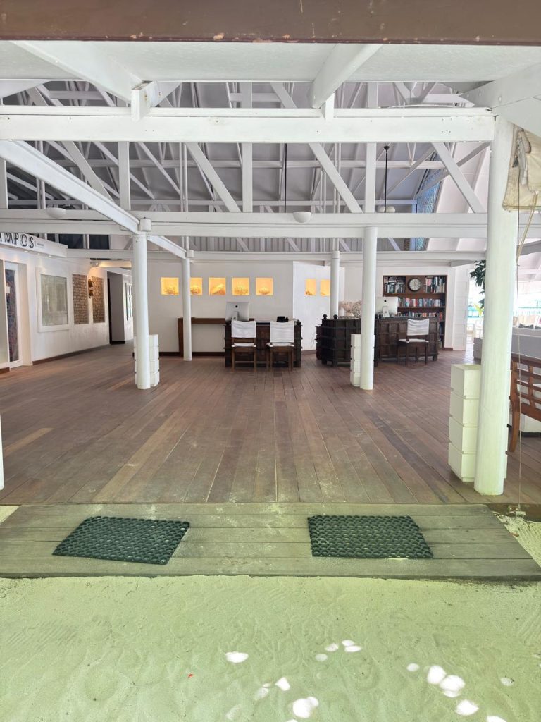 Open-air restaurant with white pillars and wooden floors, featuring a central dining table and bookshelf in the background. The entrance has two black mats and transitions from sand to the wooden interior.