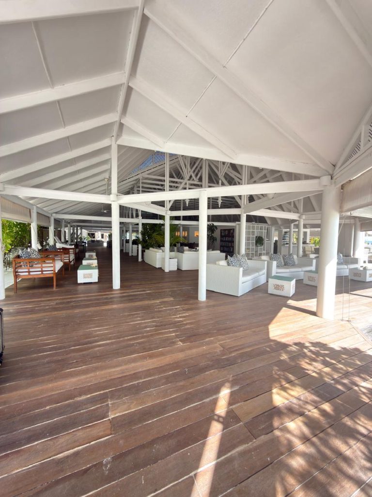 Open-air lounge with white vaulted ceiling, wooden floor, and modern white furniture. Sunlight streams in, creating shadows. Green plants and seating areas are arranged throughout the spacious, airy setting.