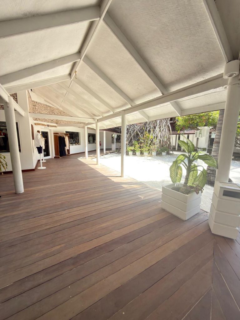 A covered wooden walkway with white pillars leads to a building with mannequins in the window. A large potted plant sits on the right, and trees and sunlight are visible in the background.