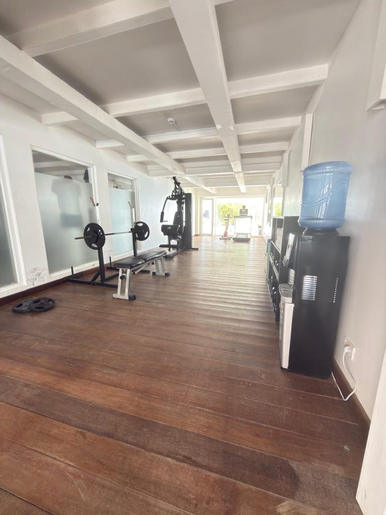 A home gym with wooden floors featuring a weight bench, gym equipment, and a water dispenser, with large windows letting in natural light.