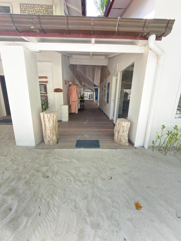 A sandy pathway leads to a covered, wooden-floored entrance of a white building with two tree trunk planters and hanging clothes on display inside. The area is bright and beach-like, with plants and natural light.