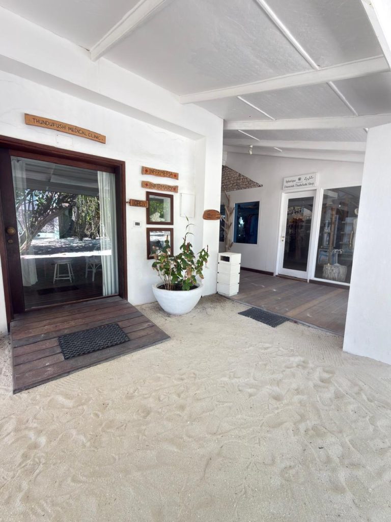 A sandy entrance area leads to two adjacent doorways of white buildings. One door has wooden signs and framed photos above, next to a large potted plant; the other has glass doors and a small mat on the sandy floor.