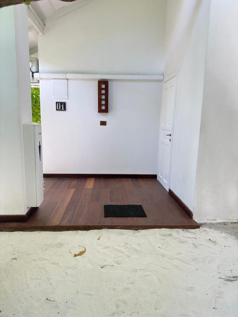 Entrance to a white building with wooden flooring, a black doormat, and a door numbered 01. The area in front of the entrance is covered with white sand.