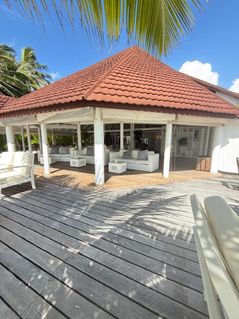 An outdoor lounge area with white cushioned seating under a red-tiled roof gazebo, surrounded by palm trees and wooden decking, in bright, sunny weather.