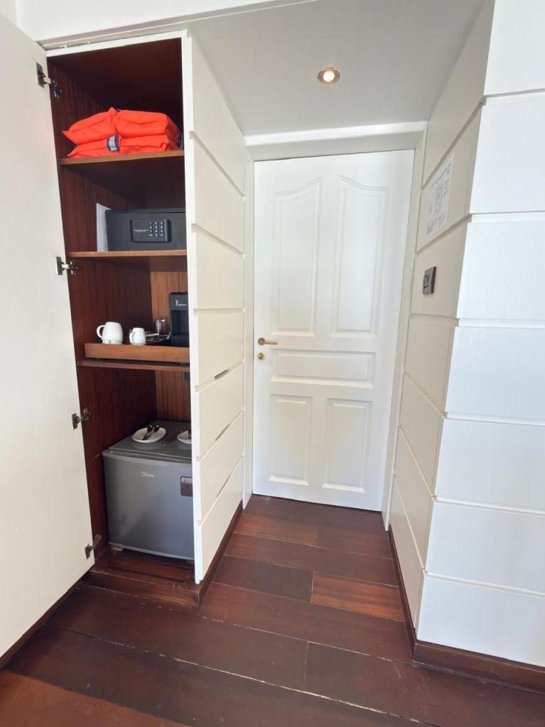 A hotel room closet with open doors reveals shelves holding a safe, two orange life jackets, a tray with cups and a kettle, and a mini fridge. A closed white door is in the center. The floor is dark wood.