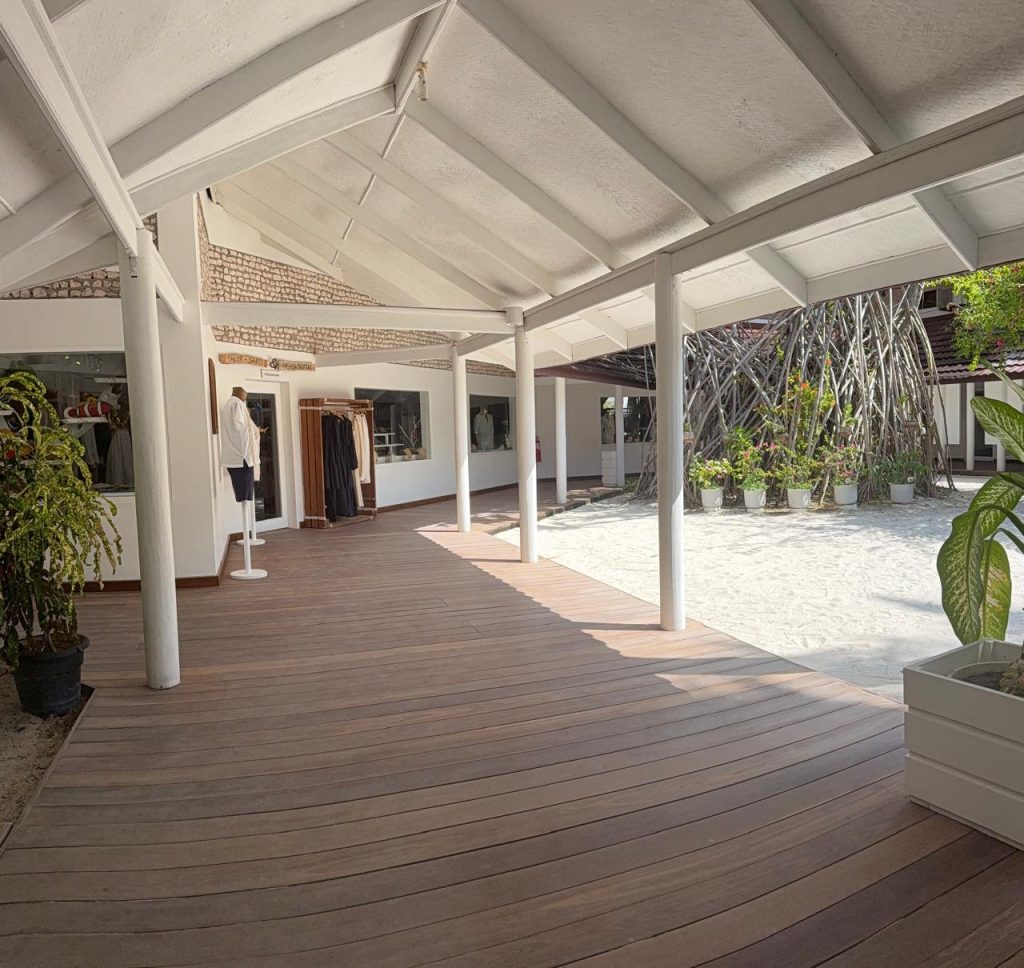 A wooden walkway with white pillars leads to a boutique with clothing displays. Potted plants line the path, and sunlight fills the open, airy courtyard with sand and a decorative wooden structure in the background.