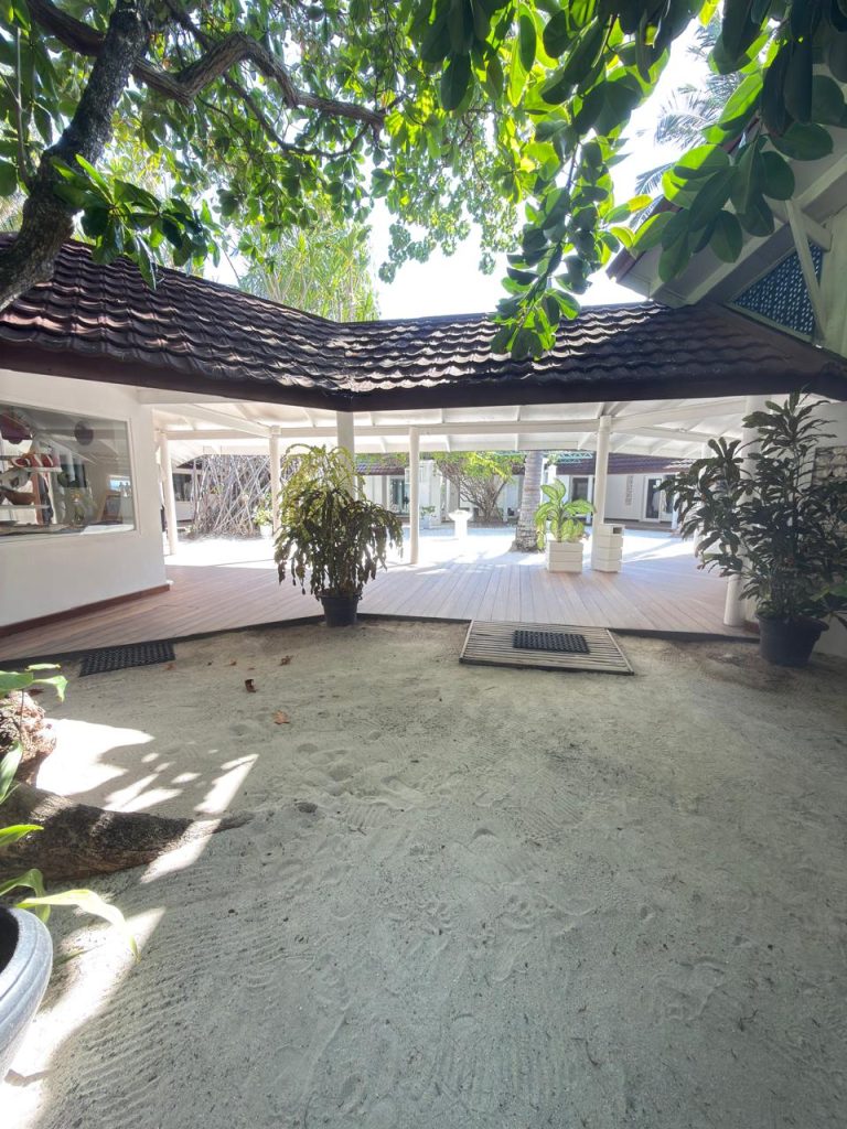 A shaded outdoor pavilion with a wooden deck, potted plants, and sand underfoot, surrounded by leafy trees and white buildings with dark brown roofs. Sunlight filters through the foliage.