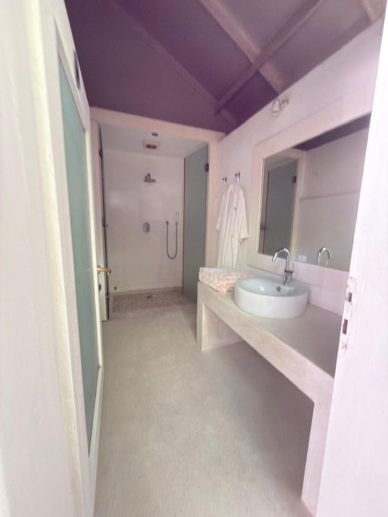 A bright, minimalist bathroom with a walk-in shower, a white sink on a counter, a large mirror, and two white robes hanging on the wall. The space has a clean, modern design with neutral tones.