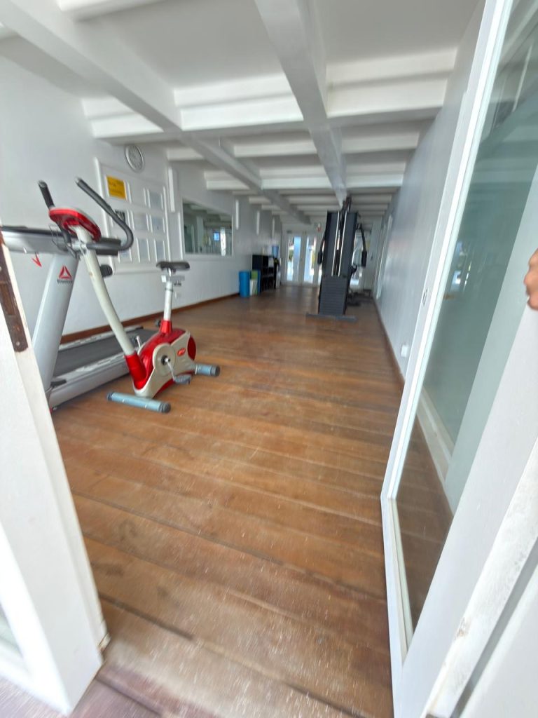 A gym room with wooden floors, featuring a red and gray exercise bike in the foreground and additional gym equipment lined up along the glass wall in the background.