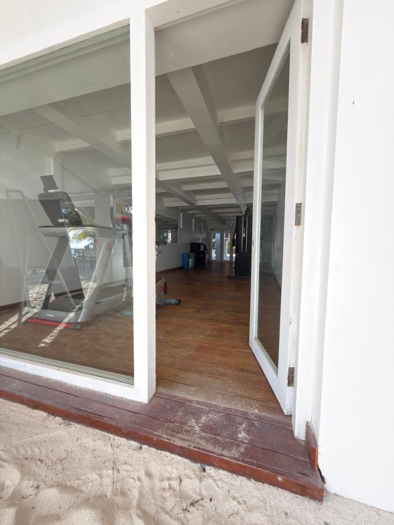 Open glass door leading from sandy outdoors into a modern gym with wooden floors, fitness equipment visible inside, and white ceiling beams. Natural light enters through large windows.