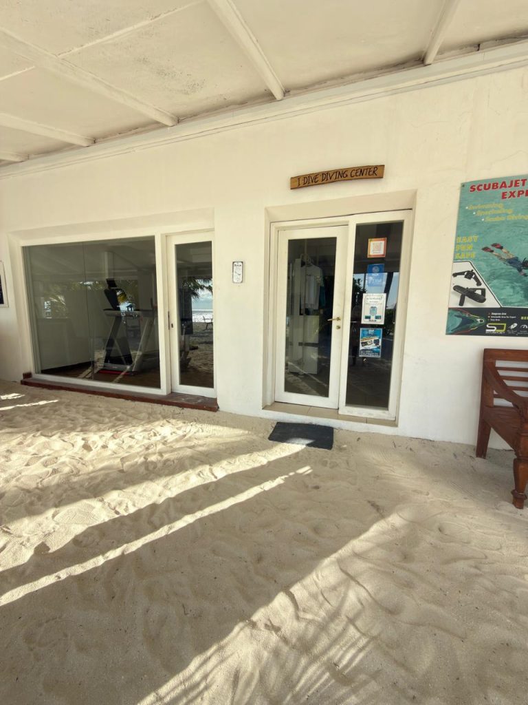 A dive center entrance with glass doors and windows, set in a white building with sand covering the floor outside. A sign above reads DIVE DIVING CENTER and a scuba diving poster is on the wall beside the door.