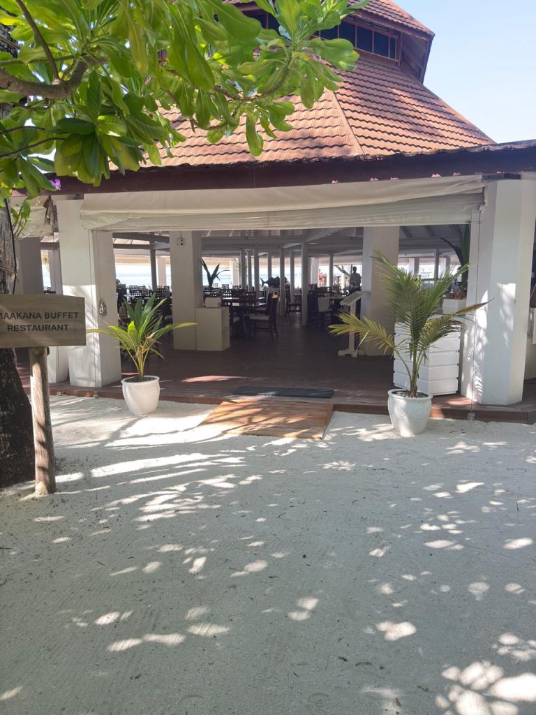 Entrance to a beachside buffet restaurant with white walls, potted plants, and a terracotta roof, seen from a sandy path shaded by leafy green trees. A sign reads JAKANA BUFFET RESTAURANT.
