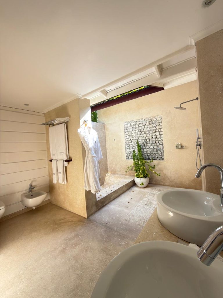 A modern bathroom with light beige walls and floor, double sinks, a bidet, towels, and an open-air shower area with natural light, plants, and a stone accent wall. A robe hangs near the shower.