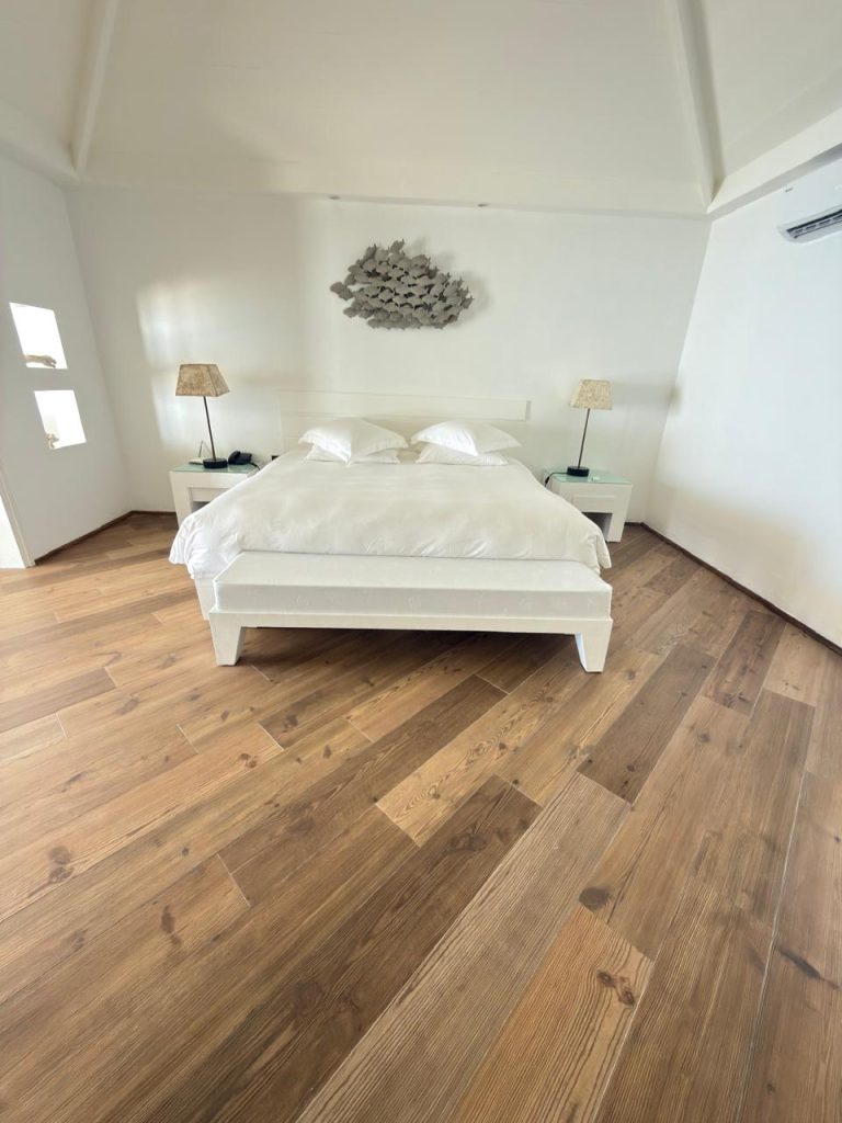 A minimalist bedroom with white walls and ceiling, a white bed with two pillows, two bedside tables with lamps, wooden flooring, and a decorative wall piece above the bed.