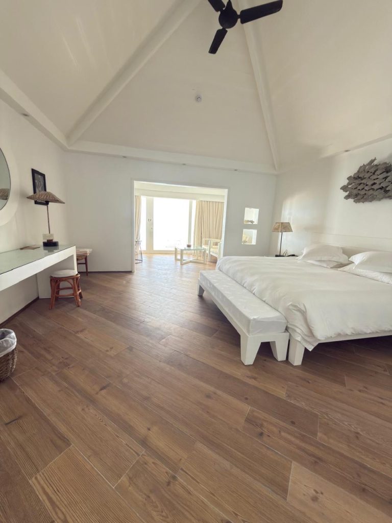A spacious, modern bedroom with white walls, a large bed, wooden flooring, a ceiling fan, desk with a mirror, and sliding doors leading to a bright balcony with outdoor seating.
