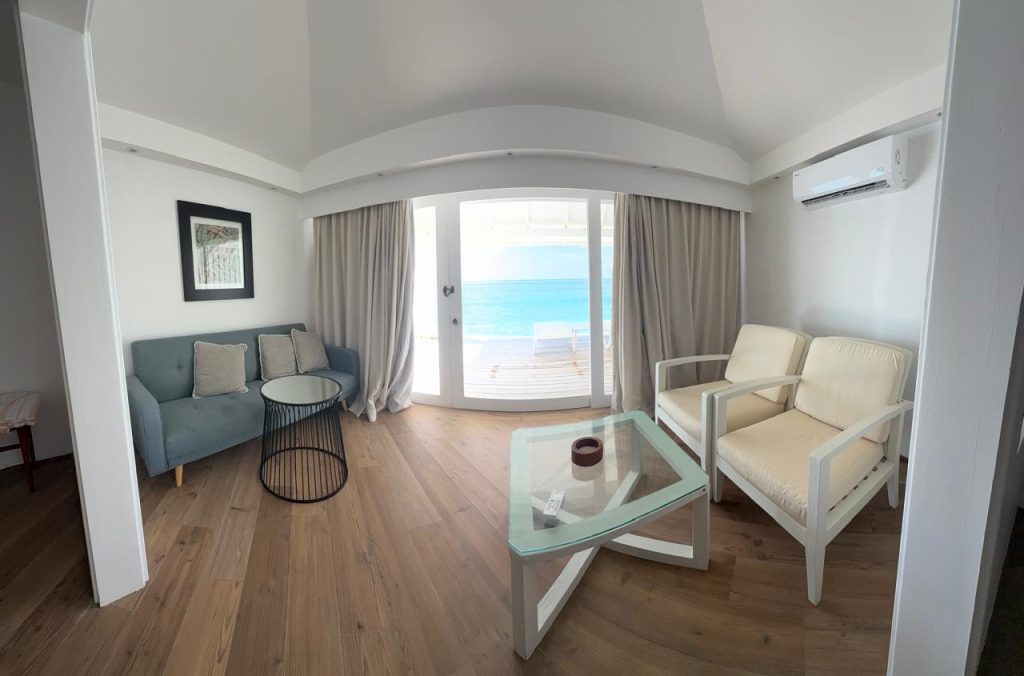 Bright, modern living room with wood floors, a glass coffee table, a blue sofa, two cream chairs, and large glass doors opening to a deck with an ocean view. Curtains are partially open, and artwork hangs on the wall.